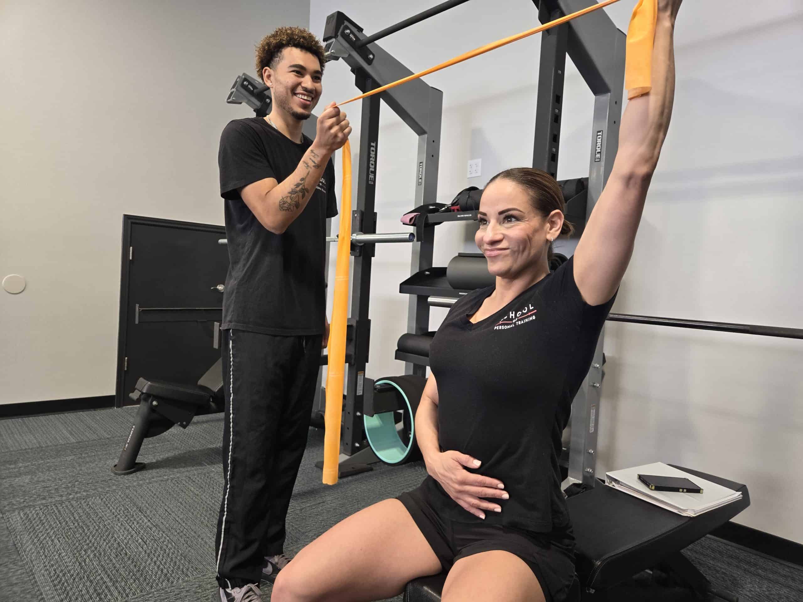 Personal training student assisting a client with resistance band exercises, demonstrating effective coaching techniques and personalized fitness program design for diverse client needs.