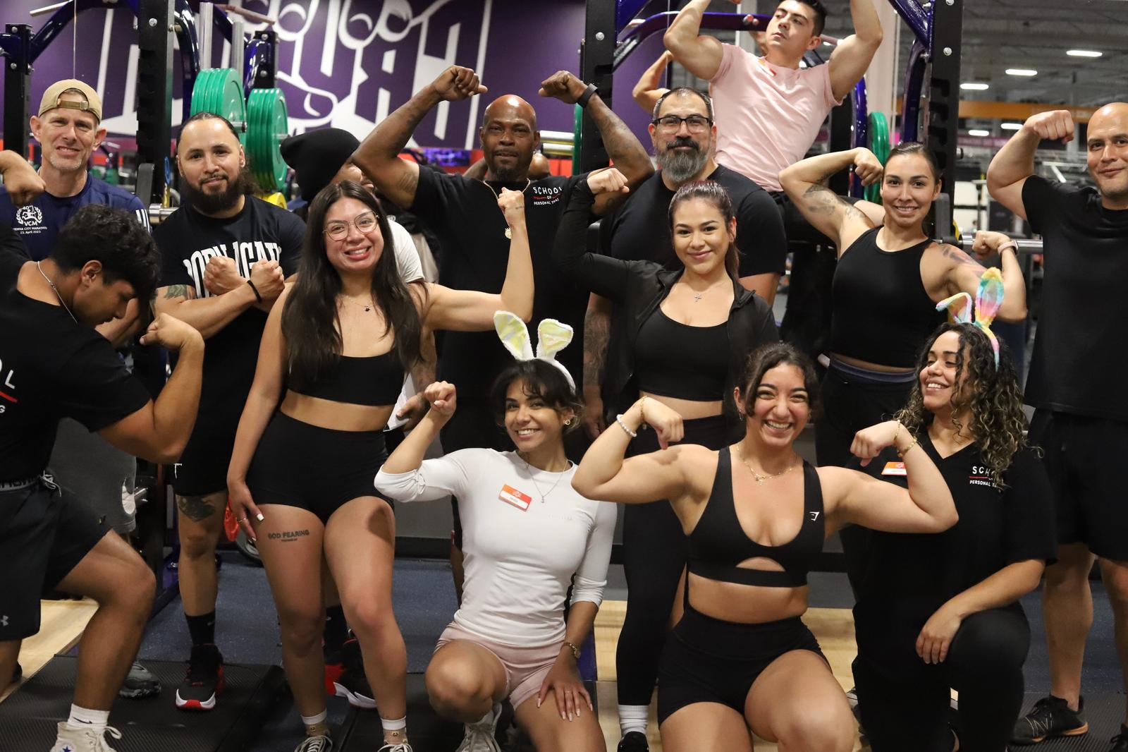 Diverse group of energized students and instructors at the School of Personal Training at Crunch Fitness in Castle Hills, Texas, flexing muscles and celebrating their fitness achievements and education.