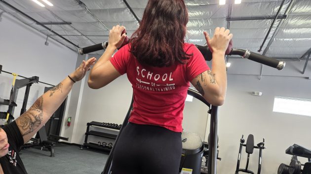 Female student at the School of Personal Training performing a machine exercise under the guidance of an instructor, emphasizing practical application and supervised fitness training.